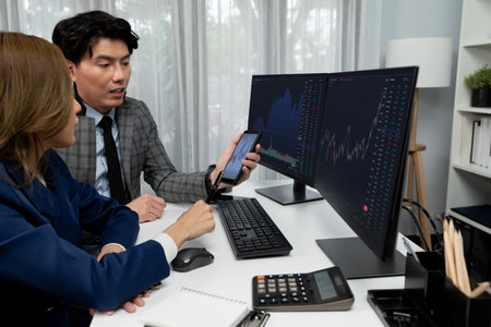 Smiling businessman showing dynamic stock market data on mobile phone to woman writing to memo for...