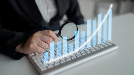 A business professional examines data trends with a magnifying glass over a keyboard, symbolizing...
