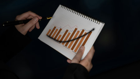 A person closely analyzes a financial bar graph in a notebook, using a pen to highlight important...