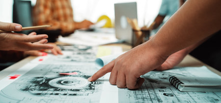 A diverse group of professionals engages in a productive brainstorming session, analyzing blueprints and project plans while using digital tools in a dynamic workspace.の写真素材