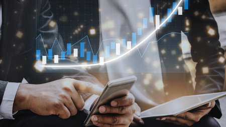 Two business professionals engage with a smartphone and tablet while examining financial charts and...