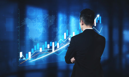 A businessman stands with arms crossed while analyzing financial data presented on a digital screen,...