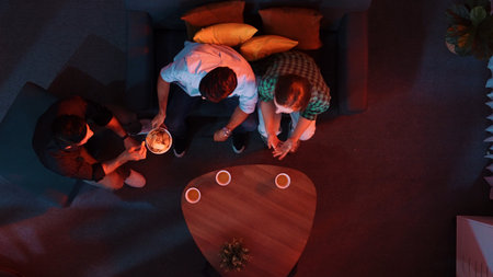 Top aerial view of caucasian father and mother watching movie while eating snack with friend. Happy family with colleague looking at television while dad holds snack bowl at living room.の写真素材