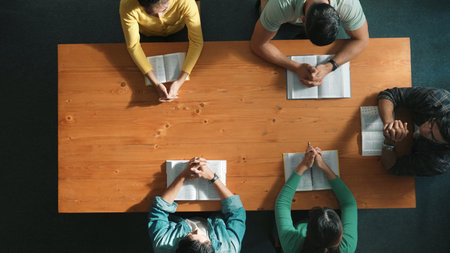 Top down aerial view of a group of people holding hands on a holy book and praying while sitting at a meeting table. Group of people with hands folded in prayer, demonstrating faith, spirituality, and religion. Symposium.の写真素材