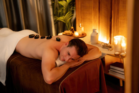 Hot stone massage at spa salon in luxury resort with warm candle light, blissful man customer enjoying spa basalt stone massage glide over body with soothing warmth. Quiescentの写真素材