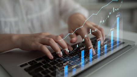 A focused individual types on a laptop while financial graphs and data trends overlay the image. The...