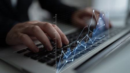 A focused view of hands typing on a laptop keyboard with financial graphs and data displayed,...