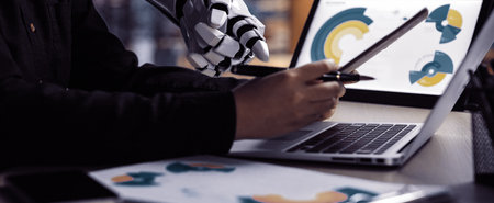 A robotic hand engages with a human partner, highlighting a collaborative effort on data analysis displayed on a laptop, showcasing modern workplace synergy.の写真素材