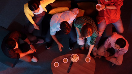 Diverse friends cheering together while happy man holding snack bowl and remote at dark living room. People watching movie and spending time together while eating snack.の写真素材