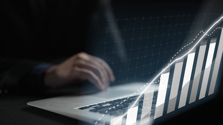 A close-up of hands typing on a laptop with a digital graph illustrating growth trends, set in a sleek office environment, representing data analysis and financial progress.の写真素材