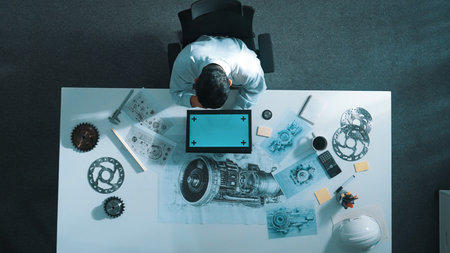 Top view of skilled engineer writing in paper while looking at tablet display green screen with turbine engine sketch design and metal gear and architectural equipment scatter around.の写真素材