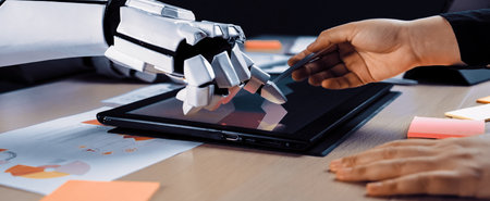 A dynamic interaction between a human hand and a robotic arm using a touchscreen tablet in a modern office setting, showcasing innovation and creativity in teamwork.の写真素材