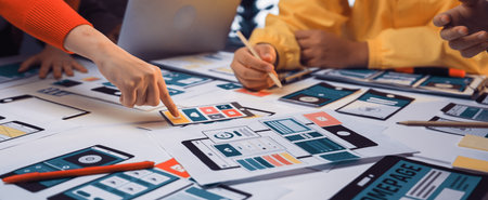 A diverse team engages in a collaborative session, strategizing the design of a mobile application with colorful wireframes and various prototypes laid out on the table.の写真素材