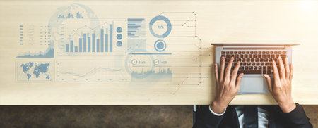 A pair of hands works on a laptop on a wooden desk. Digital graphs and charts illustrate business...