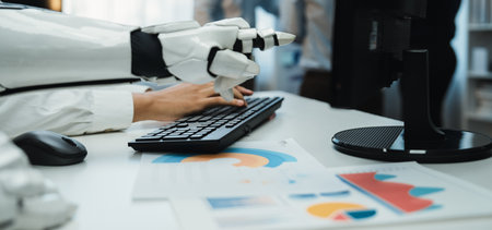 A robotic hand skillfully types on a keyboard in an office setting, surrounded by analytical charts and business documents, emphasizing technological advancement in workplaces.の写真素材
