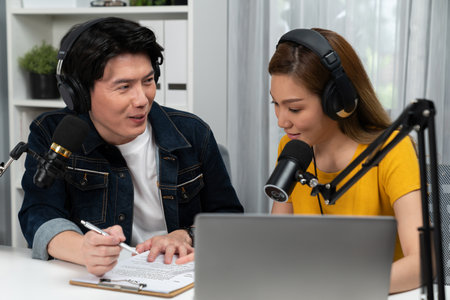 Smiling radio influencer hosts wearing headphones with channel social media online live streaming with talking topic on script note and laptop to listeners at morning time at studio record.の写真素材