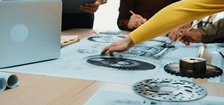 A diverse team collaborates around a table filled with technical drawings and design tools. Focus on hands working on engineering concepts in a modern office setting.の写真素材