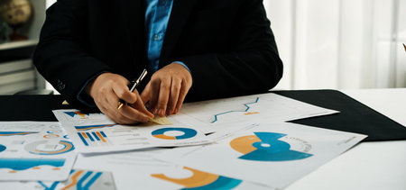 A business professional focuses intently on various financial reports, using a pen while surrounded by colorful charts and documents in a bright office space.の写真素材