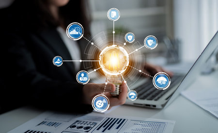 A business professional engages with a laptop while holding a light bulb, surrounded by digital icons symbolizing technology, finance, and innovation in a modern office.の写真素材