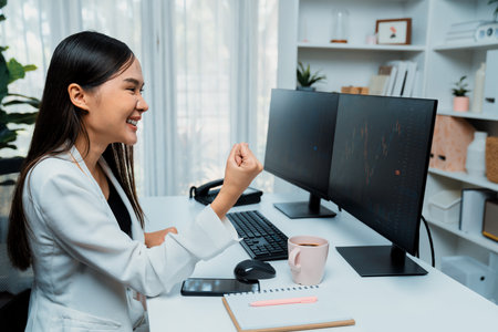 Successful young Asian businesswoman earning highest profit with fist up in exchange stock market on dynamic business graph on pc screens at modern office, analyzing financial technology. Stratagem.の写真素材
