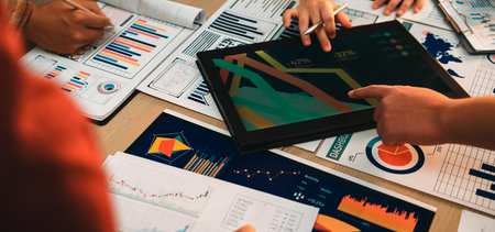 A dynamic team engaged in a business meeting, analyzing data with printed reports and a tablet. Multiple hands highlight graphs and strategies for effective decision-making.の写真素材
