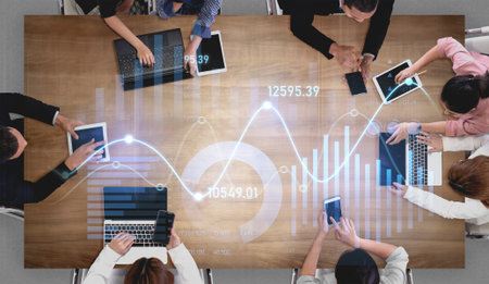 A diverse business team collaborates at a modern office table, analyzing financial data and graphical insights using various digital devices and tools.の写真素材