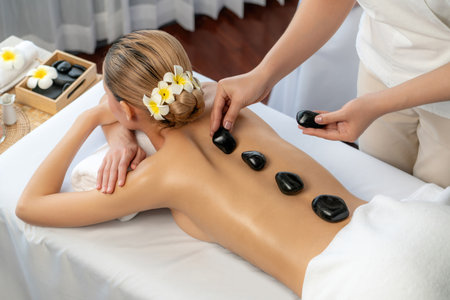 Hot stone massage at spa salon in luxury resort with day light serenity ambient, blissful woman customer enjoying spa basalt stone massage glide over body with soothing warmth. Quiescentの写真素材