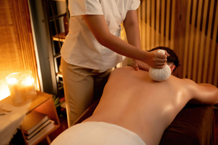 Hot herbal ball spa massage body treatment, masseur gently compresses herb bag on man body. Tranquil and serenity of aromatherapy recreation in warm lighting of candles at spa salon. Quiescentの写真素材