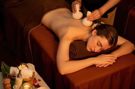 Hot herbal ball spa massage body treatment, masseur gently compresses herb bag on woman body. Tranquil and serenity of aromatherapy recreation in warm lighting of candles at spa salon. Quiescentの写真素材