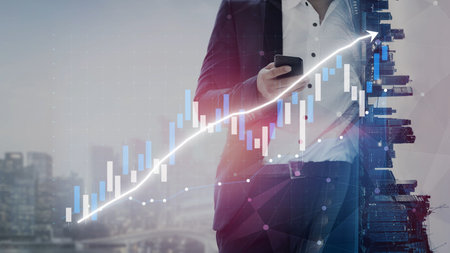 A businessman is engaged with a smartphone against a backdrop of a rising financial chart, highlighting growth and success in modern business environments.の写真素材