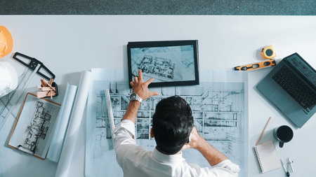 Top down view of skilled engineer talking to manager by using mobile phone while raised hand to celebrate successful project at meeting table with safety helmet, laptop and equipment.の写真素材