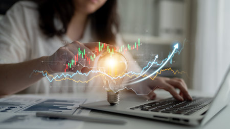 A businesswoman engages with digital data on her laptop, showcasing a light bulb animation symbolizing innovation and growth in a modern office space.の写真素材