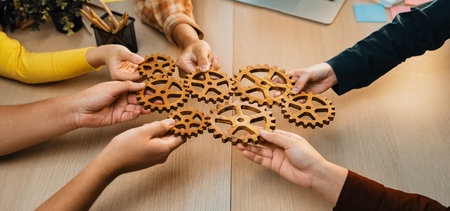 A group of diverse hands holds wooden gears in a collaborative workspace, symbolizing teamwork and innovation during a planning session in a modern office.の写真素材