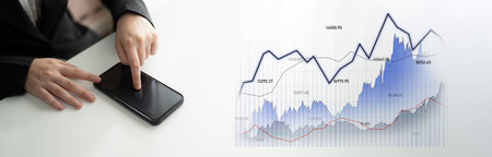 A business professional interacts with a smartphone while analyzing financial data displayed in...