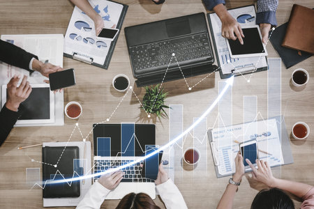 A dynamic business meeting scene showcasing professionals engaged in analysis of data trends using laptops and tablets, complemented by graphs and charts.の写真素材