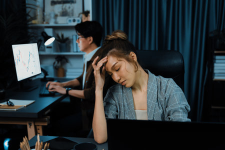 Businesswoman working on desk with stretching arm up and down manner with body health ache of strain overwork while coworker trading stock market on website on pc at late over night time.の写真素材