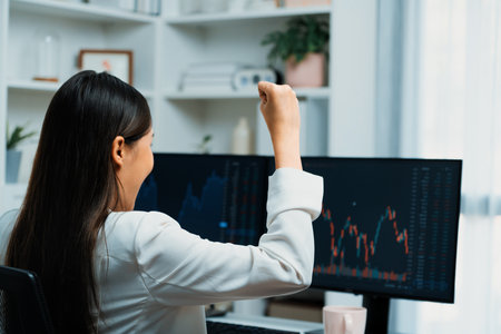 Successful young Asian businesswoman earning highest profit with fist up in exchange stock market on dynamic business graph on pc screens at modern office, analyzing financial technology. Stratagem.の写真素材
