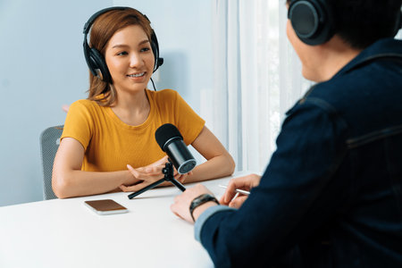 Smiling radio influencer host wearing headphone interviewing woman speaker sharing information to listeners on social media online website live streaming channel at morning at studio record.の写真素材