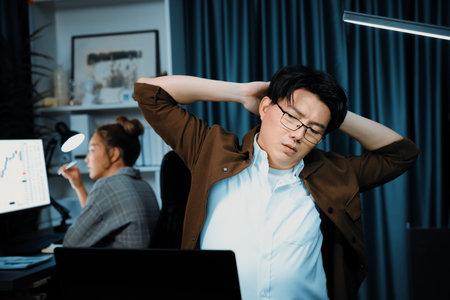 Sales manager working on desk with stretching body with arm ache at over late night time at modern home office with secretary sitting back investing dynamic stock market on project database.の写真素材