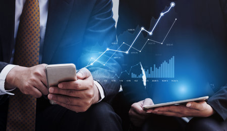Two business professionals engage with financial data on mobile devices while a growth chart...