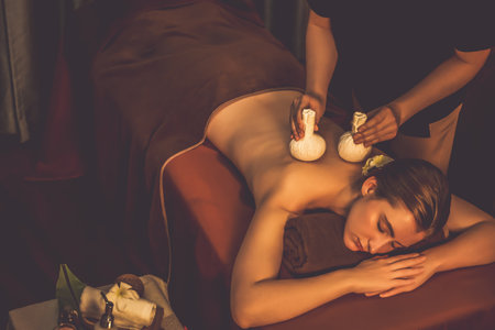 Hot herbal ball spa massage body treatment, masseur gently compresses herb bag on woman body. Tranquil and serenity of aromatherapy recreation in warm lighting of candles at spa salon. Quiescentの写真素材