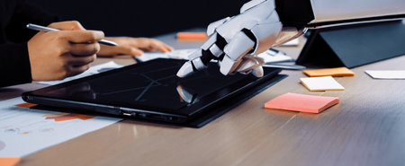 A human hand collaborates with a robotic hand on a touchscreen tablet in a modern office setting filled with colorful sticky notes and documents, symbolizing innovation.の写真素材