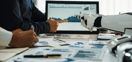 Human and robotic hands work together in a modern office, analyzing data on a computer screen filled with charts and graphs, symbolizing innovative collaboration.の写真素材