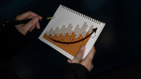 Hands holding a notepad featuring a bar graph and a rising arrow that symbolize growth and progress in business or financial analysis against a dark background.の写真素材