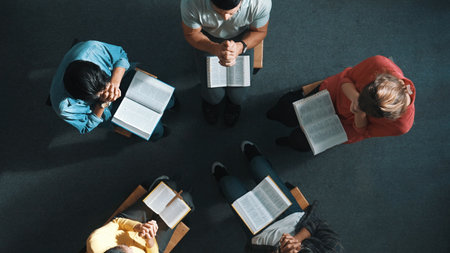 Top view of skilled diverse prayer clasping hand and praying to god while sitting at chair in circle. Top down aerial view of people folded hand while sitting with opened bible on laps. Symposium.の写真素材