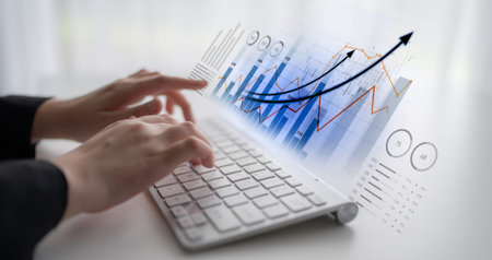 A professional setting showing hands typing on a laptop with digital financial graphs, charts, and...