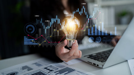 A person holding a glowing light bulb symbolizes innovation amidst financial graphs and data analysis, representing growth and strategic ideas in a contemporary office.の写真素材