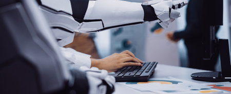 A close-up view of a futuristic robot arm typing on a computer keyboard in a modern office, showcasing advanced technology and workspace dynamics in action.の写真素材