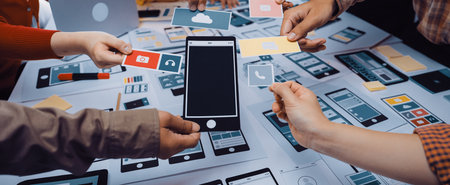 A diverse group of individuals gather around a table, collaborating on mobile application designs...