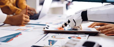 A robotic hand assists professionals in analyzing data and creating business strategies through charts and graphs in a modern office setup, showcasing technology integration.の写真素材